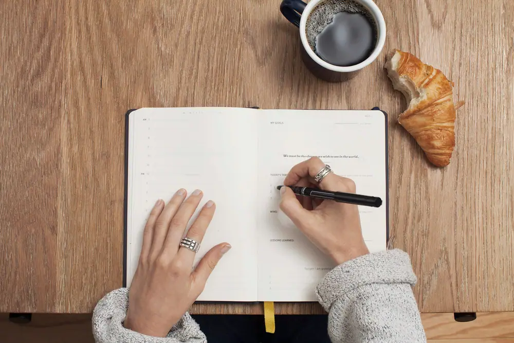 Eine Hand schreibt eine Liste in ein aufgeschlagenes Notizbuch auf einem Holztisch neben einer Kaffeetasse