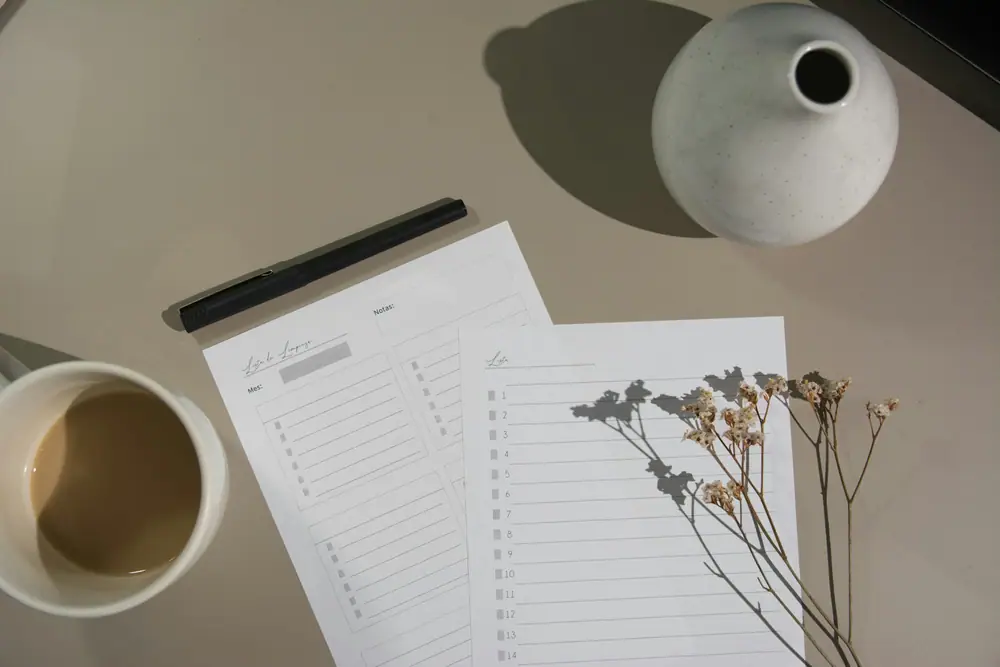 Ausgedruckte Checklisten und ein Planer auf einem Tisch neben einer Tasse Kaffee und einem getrockneten Blumenstiel