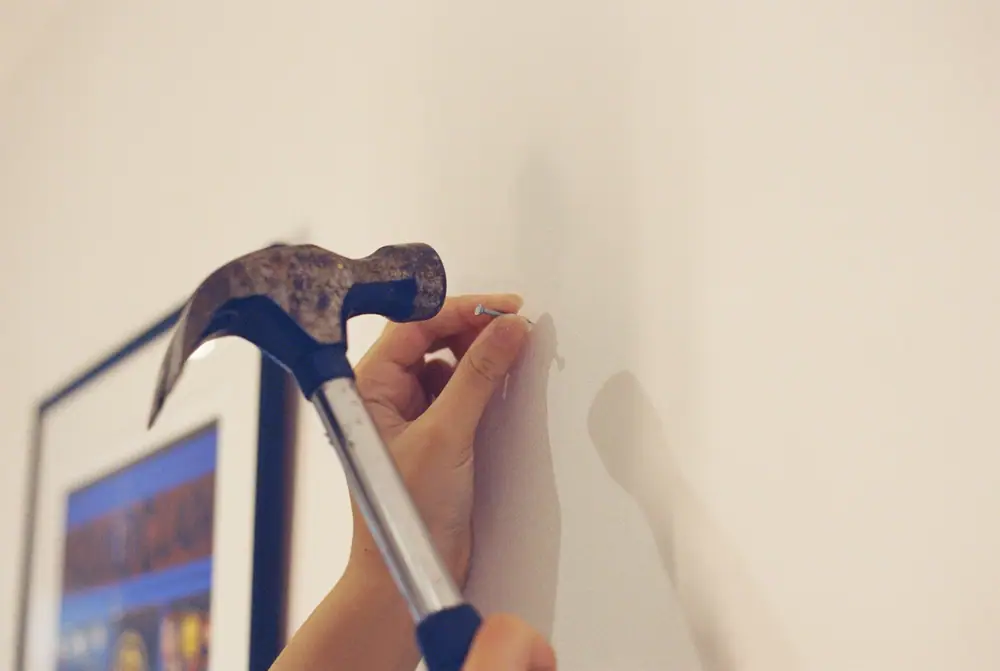 Eine Hand hält einen Nagel an eine weiße Wand, der Hammer ist zum Einschlagen erhoben